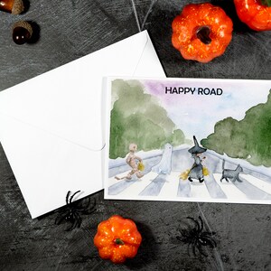 May include: A watercolor painting of a Halloween-themed scene with a skeleton, ghost, witch, and black cat walking across a crosswalk. The text "HAPPY ROAD" is written at the top of the painting.
