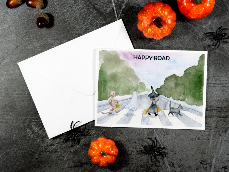 May include: A watercolor painting of a Halloween-themed scene with a skeleton, ghost, witch, and black cat walking across a crosswalk. The text "HAPPY ROAD" is written at the top of the painting.