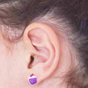 Pode incluir: Brincos de pressão em forma de cupcake roxo e branco. Os brincos são pequenos e têm uma pitada vermelha no topo.