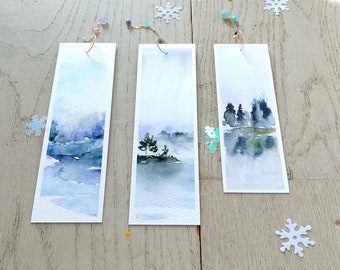 May include: Three watercolor bookmarks with wintery scenes. Each bookmark features a landscape with trees and a river, painted in shades of blue, gray, and white. The bookmarks are attached to gold chains with beads.