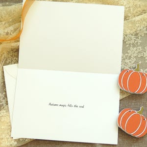 May include: A blank card with a white envelope and two orange pumpkin decorations. The card has the text "Autumn magic fills the soul" written on the inside.