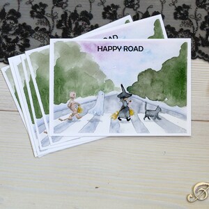 May include: A set of five watercolor postcards featuring a witch, a ghost, a mummy, and a black cat walking across a crosswalk. The text "HAPPY ROAD" is written on the top of the postcard.