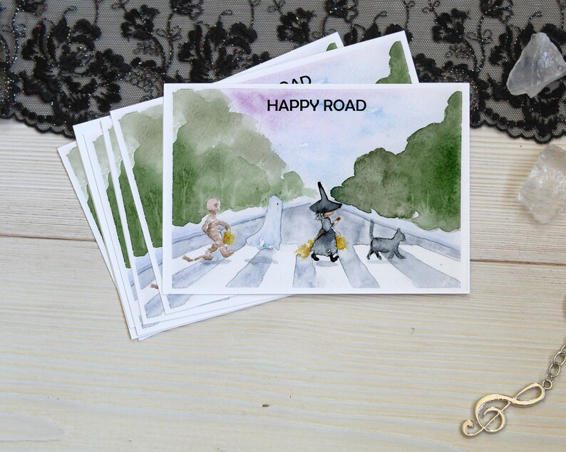 May include: A set of five watercolor postcards featuring a witch, a ghost, a mummy, and a black cat walking across a crosswalk. The text "HAPPY ROAD" is written on the top of the postcard.