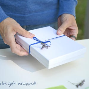 May include: A white gift box with a blue ribbon and a sprig of lavender. The box is being held by a person's hands. Can be gift wrapped!