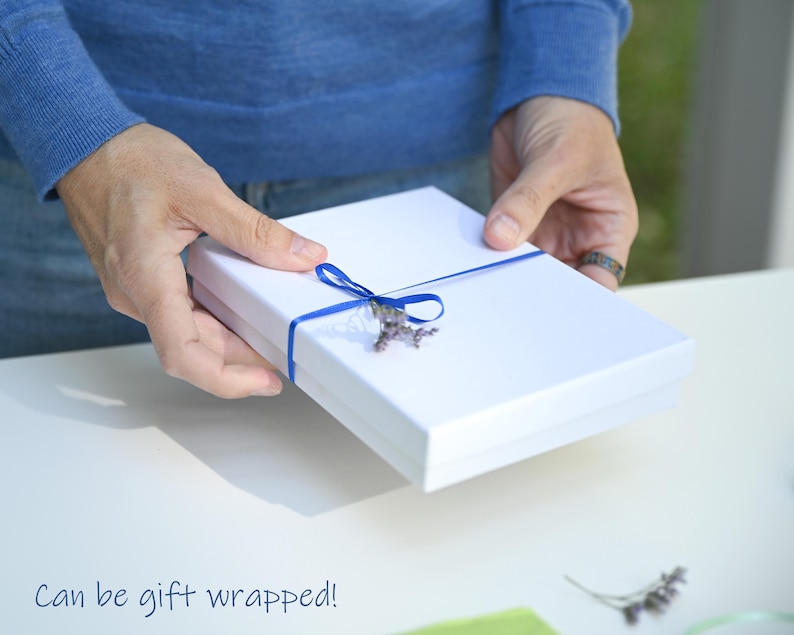 May include: A white gift box with a blue ribbon and a sprig of lavender. The box is being held by a person's hands. Can be gift wrapped!