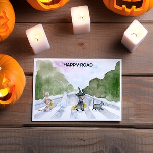 May include: A watercolor illustration of a ghost, a witch, a cat, and a mummy walking across a crosswalk. The illustration is on a white card with the text "HAPPY ROAD" at the top.