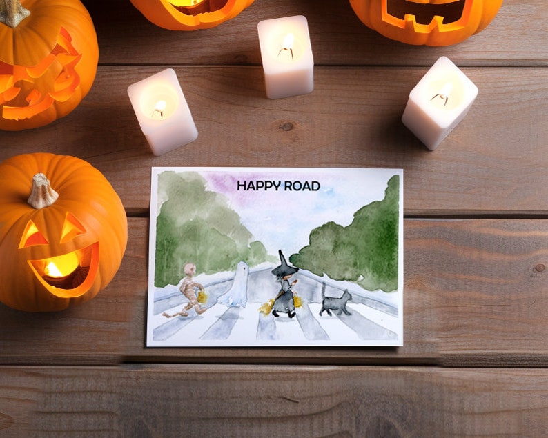 May include: A watercolor illustration of a ghost, a witch, a cat, and a mummy walking across a crosswalk. The illustration is on a white card with the text "HAPPY ROAD" at the top.