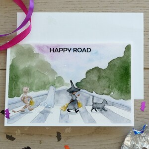 May include: A watercolor illustration of a Halloween-themed card with a witch, ghost, and a mummy walking across a crosswalk. The text "HAPPY ROAD" is written in black at the top of the card.