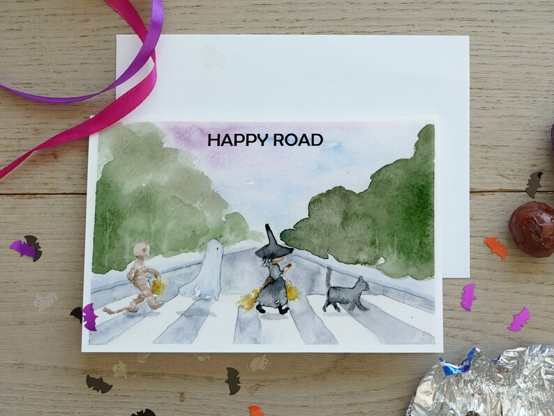 May include: A watercolor illustration of a Halloween-themed card with a witch, ghost, and a mummy walking across a crosswalk. The text "HAPPY ROAD" is written in black at the top of the card.