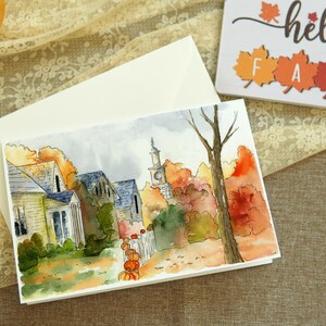 May include: Watercolour painting of a small town with houses and a church steeple in the background. The houses are painted in shades of yellow and brown, and the church steeple is painted in a light blue. The trees are painted in shades of green, orange, and red, and the ground is painted in shades of brown and green. There are pumpkins in the foreground.