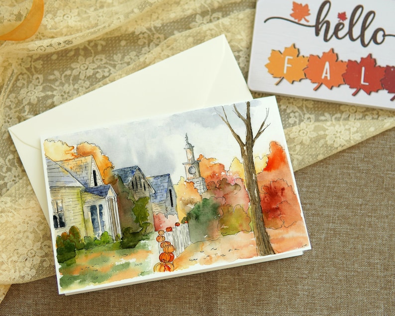 May include: Watercolour painting of a small town with houses and a church steeple in the background. The houses are painted in shades of yellow and brown, and the church steeple is painted in a light blue. The trees are painted in shades of green, orange, and red, and the ground is painted in shades of brown and green. There are pumpkins in the foreground.