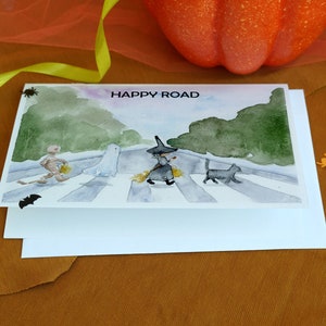 May include: A watercolor card with a Halloween theme. The card features a witch, a ghost, a mummy, and a black cat walking across a crosswalk. The text "HAPPY ROAD" is printed at the top of the card.