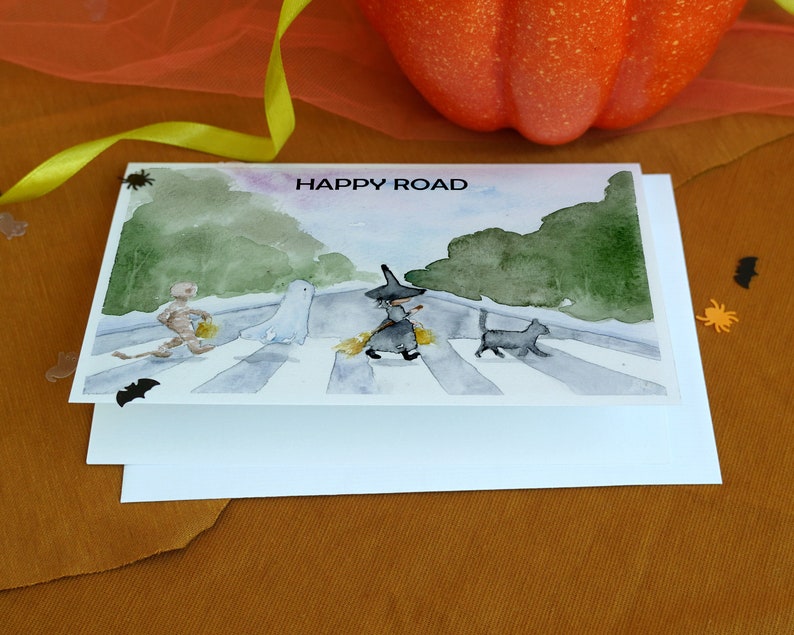 May include: A watercolor card with a Halloween theme. The card features a witch, a ghost, a mummy, and a black cat walking across a crosswalk. The text "HAPPY ROAD" is printed at the top of the card.