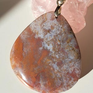 Op de afbeelding: Een druppelvormige hanger van Indiase agaat, met rode, bruine en witte patronen. De hanger heeft een zilverkleurige balein voor een ketting. De tekst "Agate indienne Padma" is zichtbaar. Een uniek sieraad.