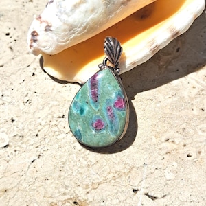 Peut inclure: Un pendentif en forme de goutte d'eau avec une bélière argentée. Le pendentif présente une pierre verte avec des inclusions rouges et bleues. Le pendentif repose sur une surface de couleur claire, avec un coquillage en arrière-plan.