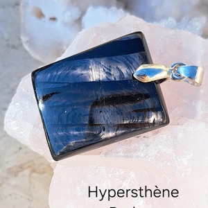 May include: A polished, dark blue hypersthene pendant with a silver-colored bail. The pendant has a rectangular shape with a slightly angled top and bottom. The text "Hypersthène Padma" is printed below the pendant.