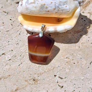 May include: A polished, geometric pendant with a silver bail. The pendant features a gradient of colors, transitioning from deep burgundy to a warm orange and a white band. The pendant is displayed near a seashell on a textured surface.