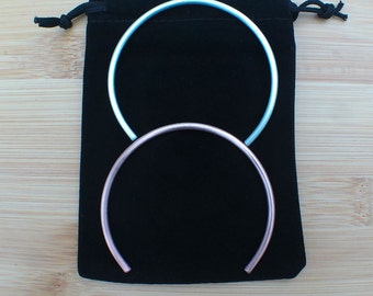 May include: Two metal bangles, one light blue and one rose gold, are displayed on a black velvet pouch with a drawstring closure.