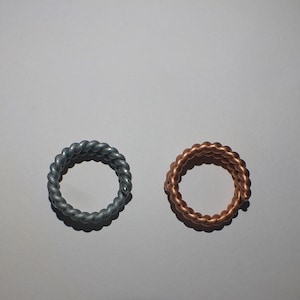 May include: Two rings with a braided rope design. One ring is silver-colored, and the other is copper-colored. The rings are circular and sit on a white surface. These rings are suitable for everyday wear.