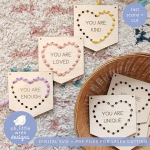 Könnte beinhalten: Holzschilder mit Laser geschnittenen, gestickten Herzmotiven. Die Schilder zeigen die Sätze „YOU ARE KIND“, „YOU ARE LOVED“, „YOU ARE ENOUGH“ und „YOU ARE UNIQUE“. Die Schilder befinden sich in einem Korb und auf einem gemusterten Teppich. Der Text „DIGITAL SVG + PDF FILES FOR LASER CUTTING“ ist ebenfalls sichtbar.