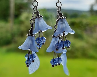 May include: A pair of silver earrings with blue flower-shaped charms and crystal accents. The earrings are hanging from a pen.