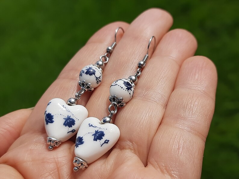 May include: A pair of dangle earrings with white heart-shaped ceramic beads with blue floral designs and silver accents.