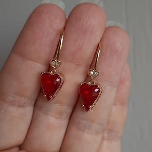 Peut inclure: Une paire de boucles d'oreilles dorées en forme de cœur avec des pierres précieuses rouges au centre. Les boucles d'oreilles ont un petit accent en forme de cœur en haut avec des pierres transparentes. Les boucles d'oreilles sont pendantes.