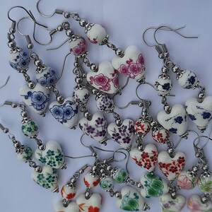 May include: A collection of white heart-shaped earrings with floral patterns in various colors, including blue, pink, red, and green. The earrings are made of ceramic and have silver-toned metal hooks.