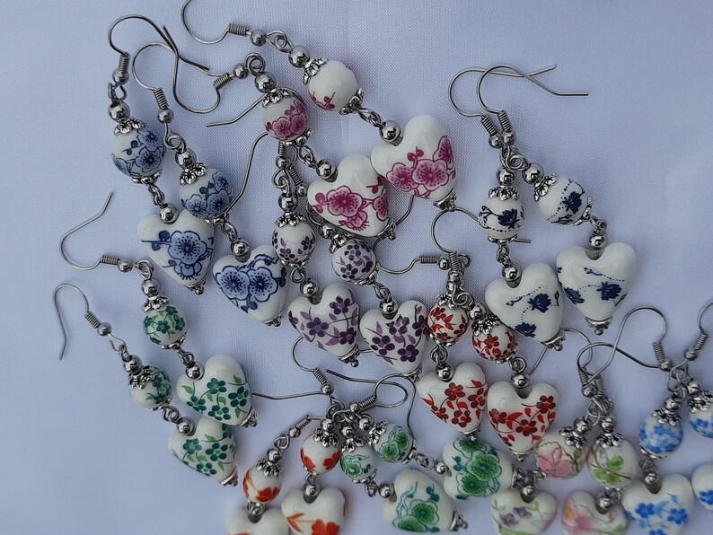 May include: A collection of white heart-shaped earrings with floral patterns in various colors, including blue, pink, red, and green. The earrings are made of ceramic and have silver-toned metal hooks.