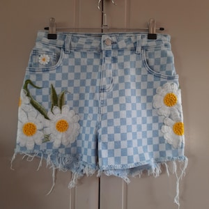 Op de afbeelding: Lichtblauwe denim shorts met een geruit patroon en geborduurde witte madeliefjes met gele centra.