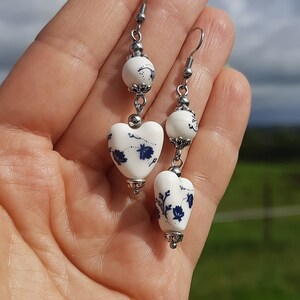 May include: A pair of white ceramic heart earrings with blue floral designs. The earrings are suspended from silver hooks and feature a white ceramic bead with a silver accent.