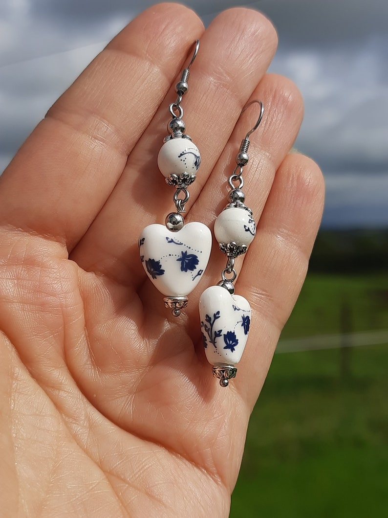 May include: A pair of white ceramic heart earrings with blue floral designs. The earrings are suspended from silver hooks and feature a white ceramic bead with a silver accent.