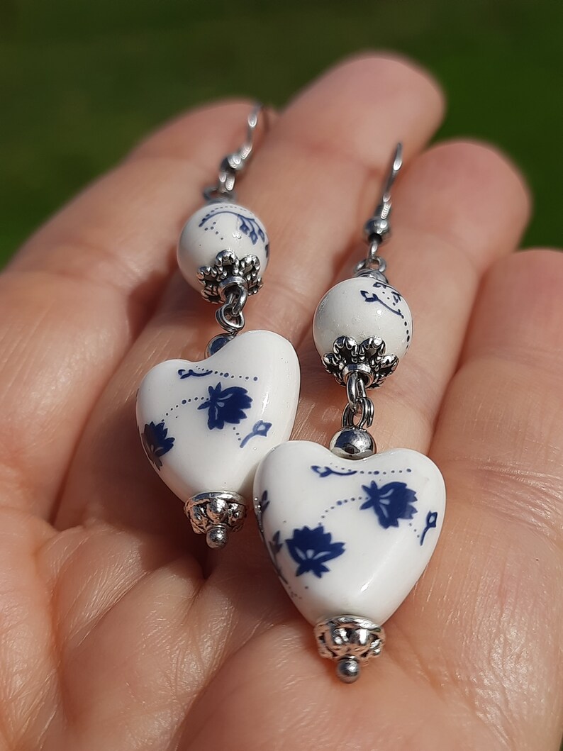 May include: A pair of dangle earrings with white heart-shaped ceramic beads with blue floral designs and silver accents.