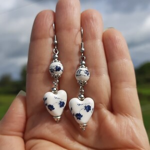 May include: A pair of dangle earrings with white heart-shaped ceramic beads with blue floral designs and silver metal accents.