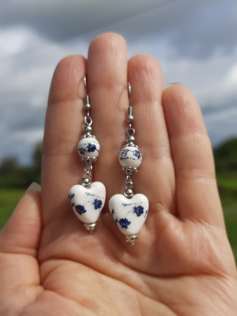 May include: A pair of dangle earrings with white heart-shaped ceramic beads with blue floral designs and silver metal accents.