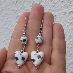 May include: A pair of dangle earrings with a white heart-shaped ceramic bead with a blue floral pattern and a smaller white round ceramic bead with a blue floral pattern. The earrings are made with silver-toned metal.