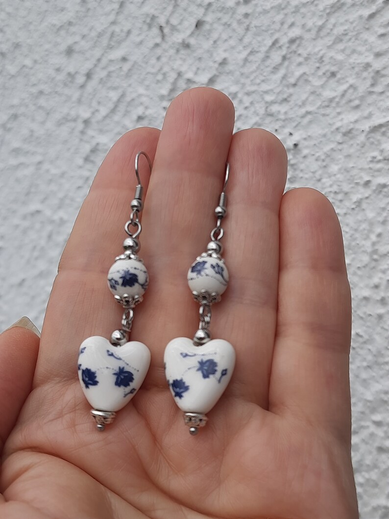 May include: A pair of dangle earrings with a white heart-shaped ceramic bead with a blue floral pattern and a smaller white round ceramic bead with a blue floral pattern. The earrings are made with silver-toned metal.