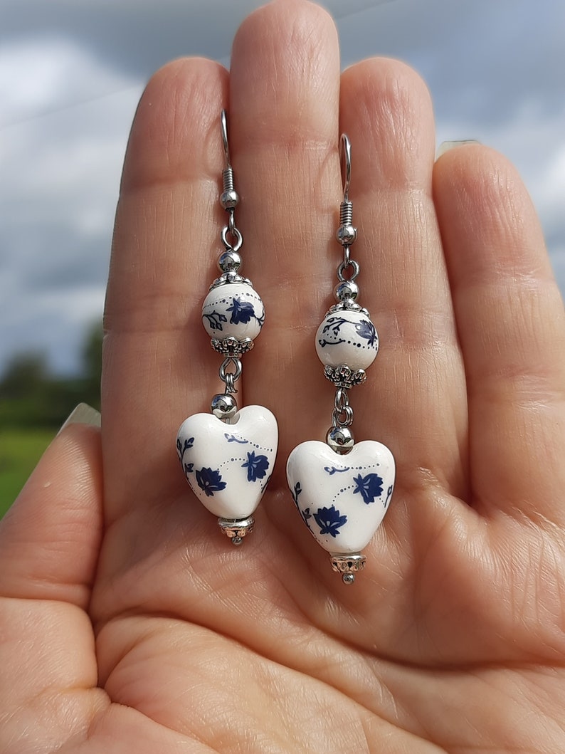 May include: A pair of silver earrings with white ceramic heart-shaped charms. The charms have a blue floral design.