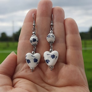 May include: A pair of silver dangle earrings with white ceramic heart-shaped charms. The charms have a blue and white floral pattern.