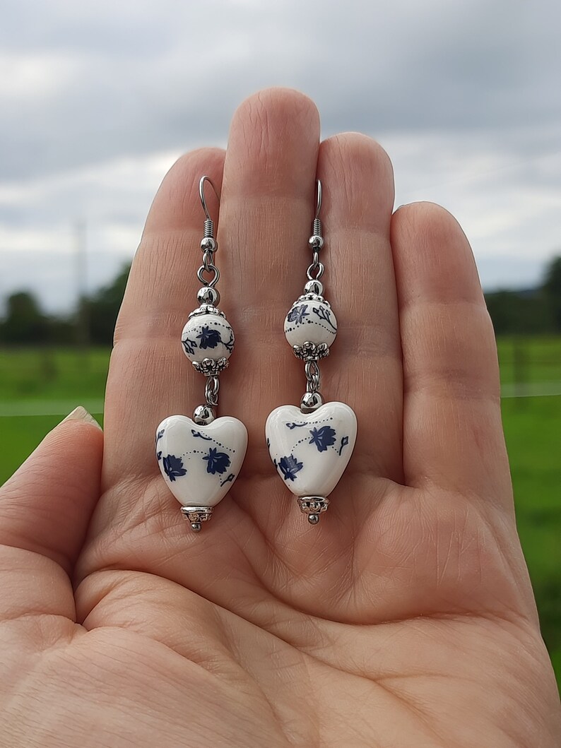 May include: A pair of silver dangle earrings with white ceramic heart-shaped charms. The charms have a blue and white floral pattern.