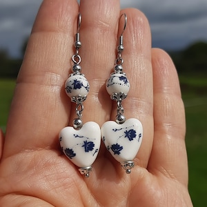 May include: A pair of dangle earrings with white porcelain heart-shaped charms with blue floral designs. The charms are attached to silver-toned beads and hooks.
