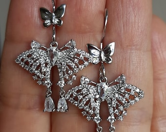 May include: Silver butterfly earrings with intricate detailing. Each earring features a small butterfly at the top, a larger butterfly design with embedded clear stones, and two teardrop-shaped clear stones dangling below.