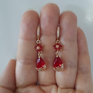 Op de afbeelding: Een paar gouden oorbellen met rode druppelvormige stenen en kleine rode bloemenaccenten. De oorbellen hebben een hoepelontwerp en zijn versierd met heldere zirkonia-stenen.