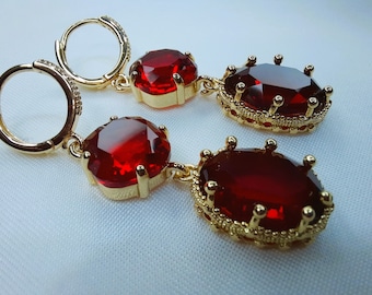 May include: A pair of gold earrings with red gemstone drops. The earrings have a hoop design and the gemstones are set in a gold crown-like setting.