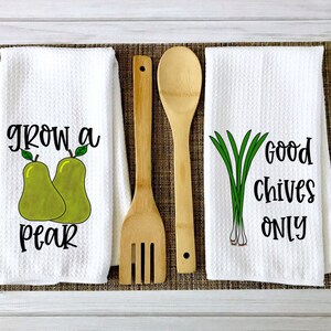 Op de afbeelding: Twee witte keukendoeken met zwarte tekst en illustraties. Een handdoek zegt "grow a pear" met twee groene peren. De andere handdoek zegt "good chives only" met een bosje groene bieslook.