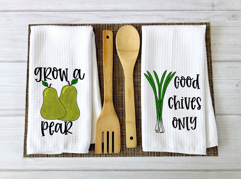 Op de afbeelding: Twee witte keukendoeken met zwarte tekst en illustraties. Een handdoek zegt "grow a pear" met twee groene peren. De andere handdoek zegt "good chives only" met een bosje groene bieslook.