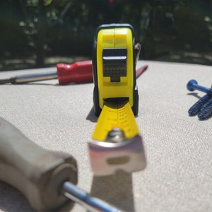 May include: A yellow measuring tape with a black and yellow case, a red and black screwdriver, and three blue screws on a white surface.