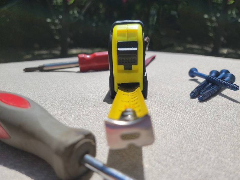 May include: A yellow measuring tape with a black and yellow case, a red and black screwdriver, and three blue screws on a white surface.