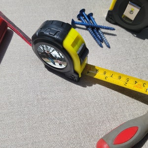 May include: A yellow and black tape measure with a reflective surface and a black and red screwdriver. The tape measure is extended to show the numbers 1 to 9. There are also blue screws on the surface.