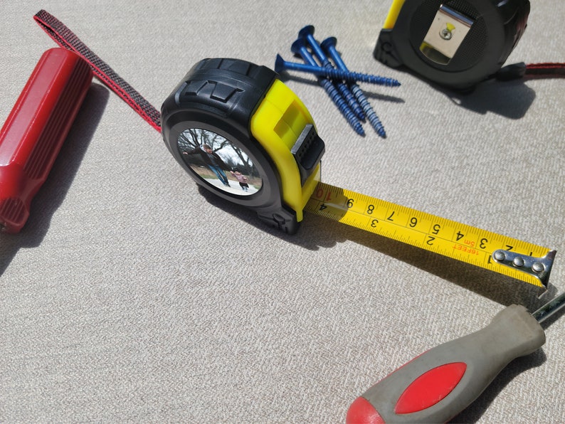 May include: A yellow and black tape measure with a reflective surface and a black and red screwdriver. The tape measure is extended to show the numbers 1 to 9. There are also blue screws on the surface.
